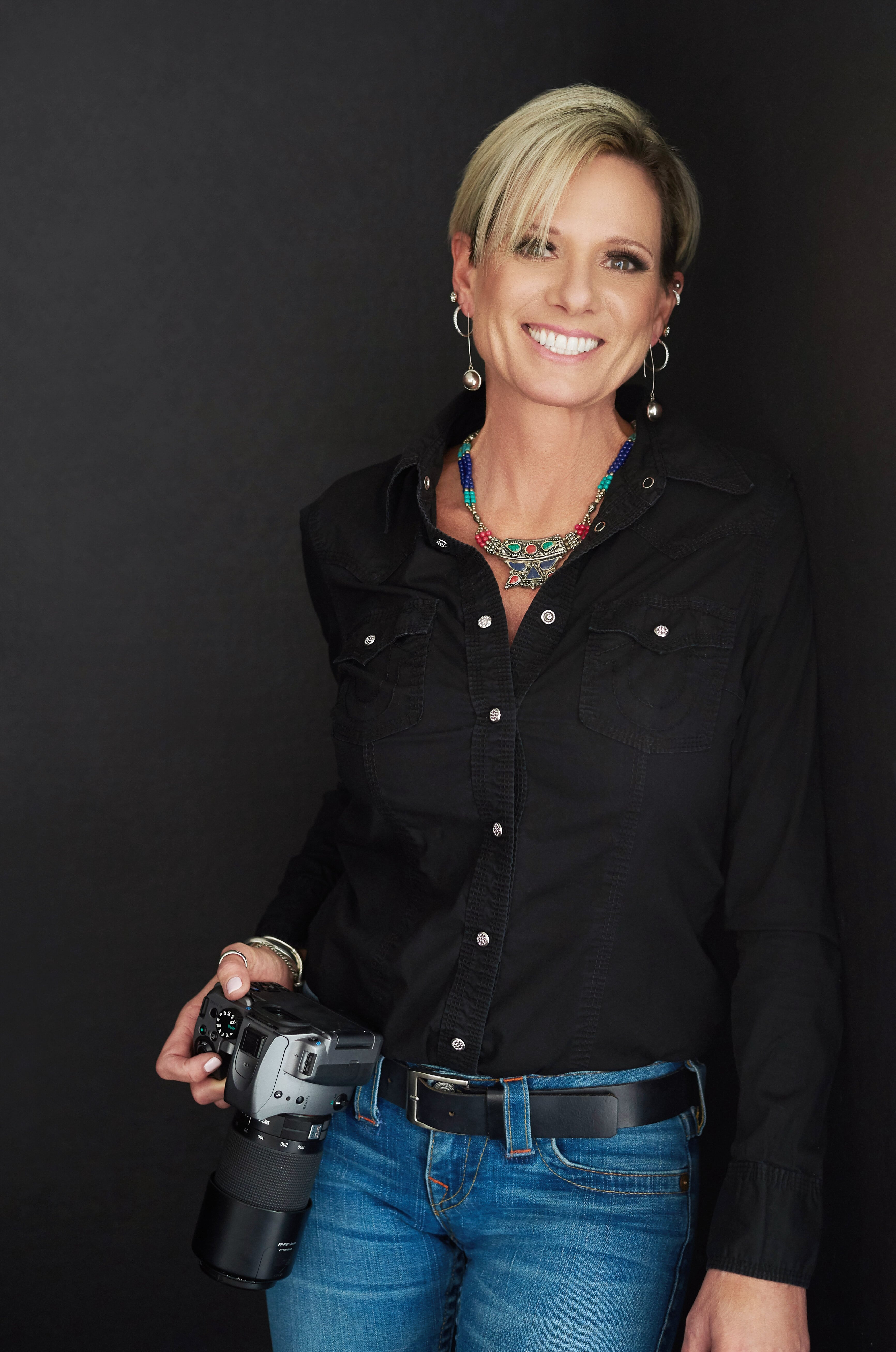 Beautiful smiling white woman photographer with short blonde hair wearing a black top, turquoise necklace, and holding a camera
