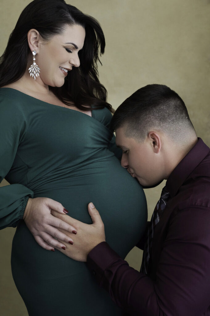 Beautiful smiling brunette pregnant woman in long green dress, husband kissing belly\