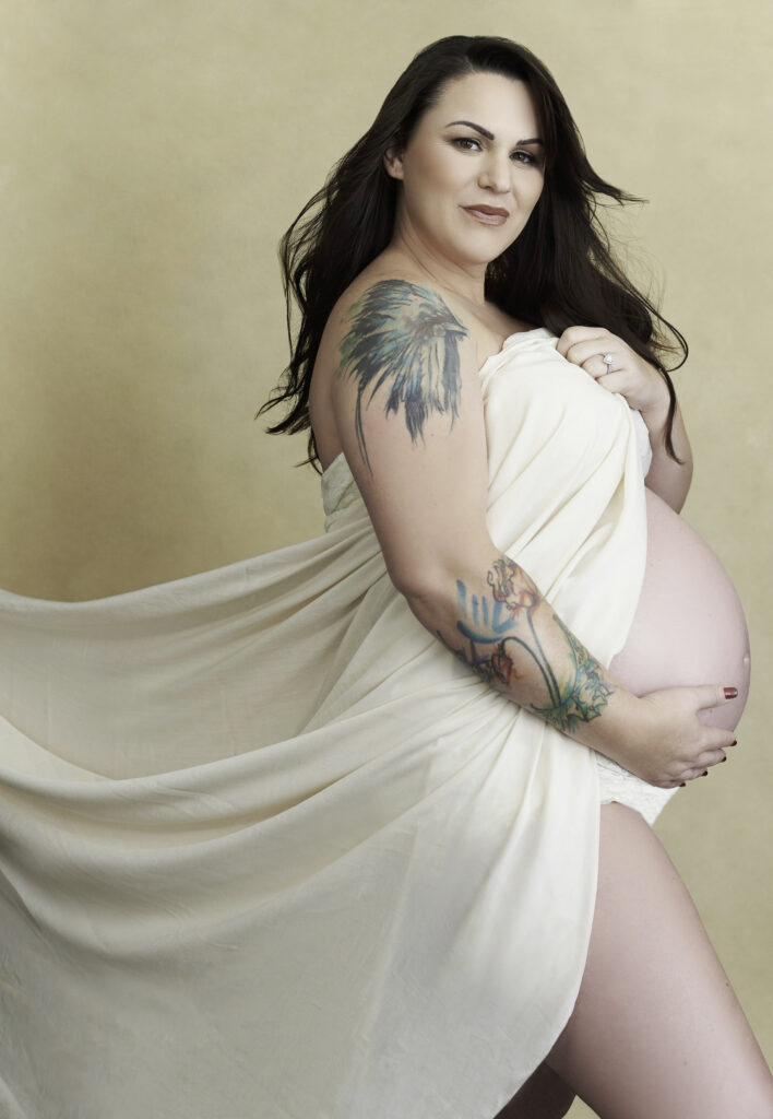 Beautiful brunette pregnant woman in flowly white dress with exposed belly and tattooes